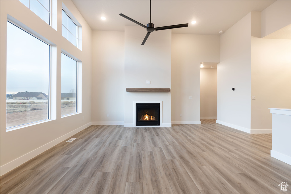Unfurnished living room featuring a lit fireplace, light wood-style flooring, baseboards, ceiling fan, and a towering ceiling