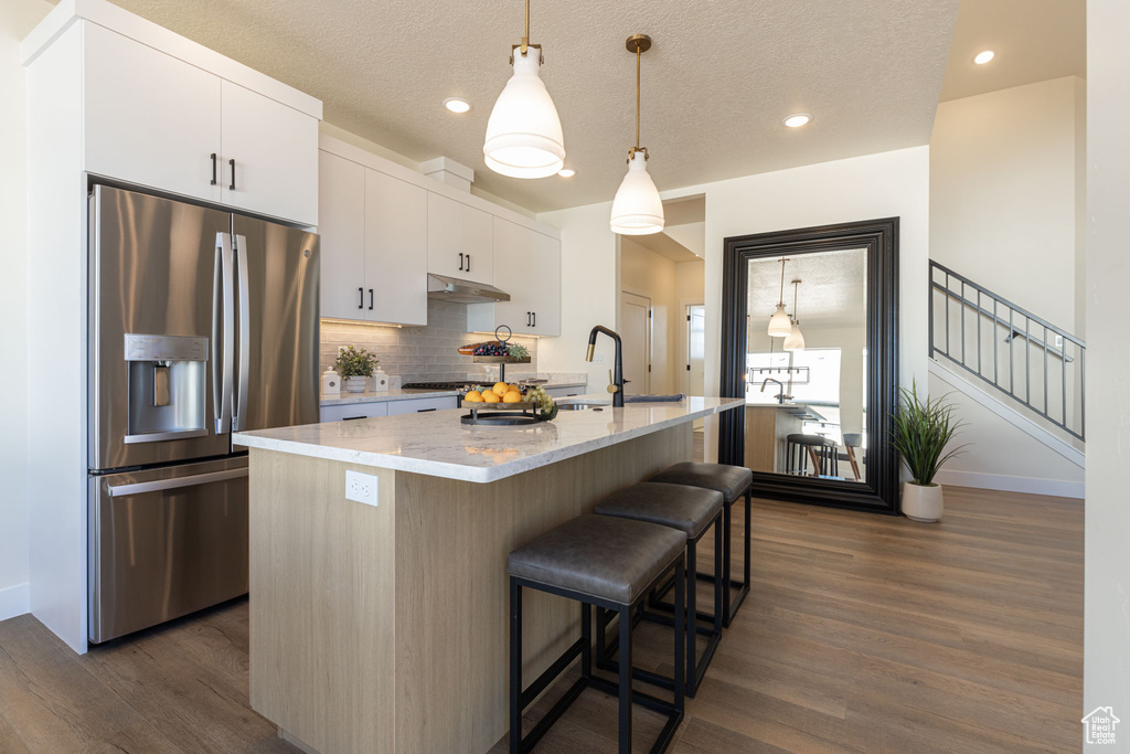 Kitchen featuring stainless steel fridge, under cabinet range hood, dark wood finished floors, decorative backsplash, and a breakfast bar