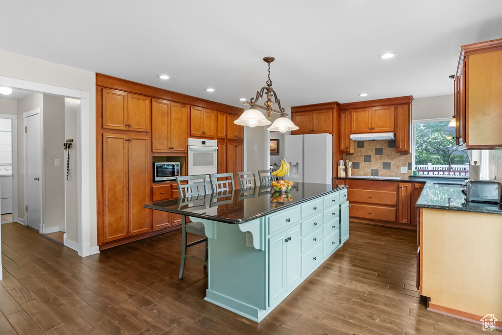 Kitchen featuring white appliances, under cabinet range hood, dark wood-style floors, a kitchen island, and brown cabinetry