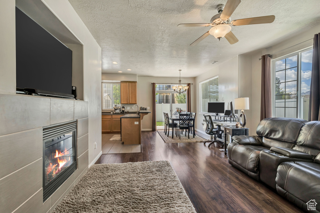 Living area with a textured ceiling, a tiled fireplace, dark wood-style flooring, a chandelier, and ceiling fan