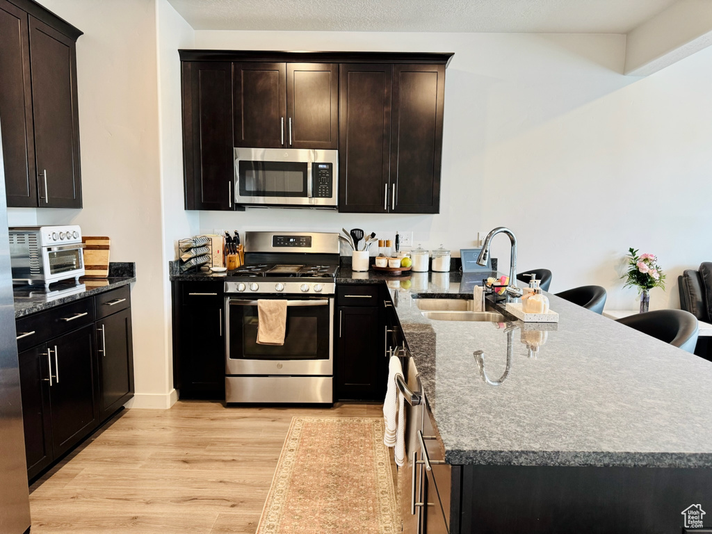 Kitchen with appliances with stainless steel finishes, a sink, a peninsula, light wood-style flooring, and dark stone counters