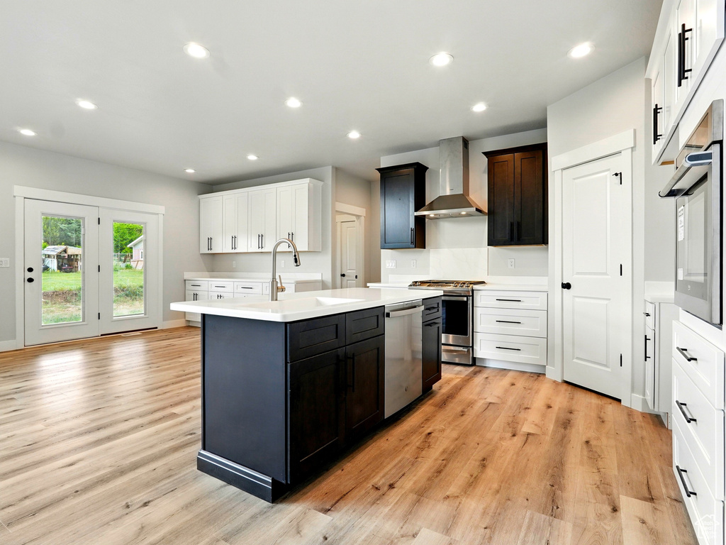 Kitchen with wall chimney range hood, stainless steel appliances, a sink, an island with sink, and light countertops