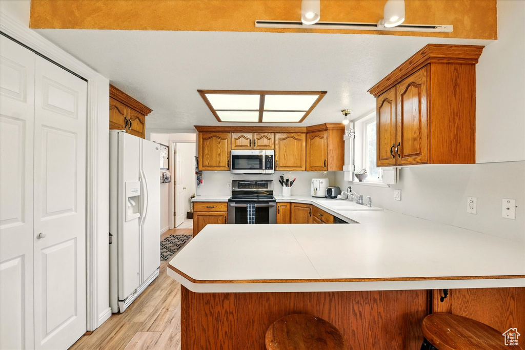 Kitchen featuring appliances with stainless steel finishes, a peninsula, a sink, brown cabinets, and light wood-style flooring