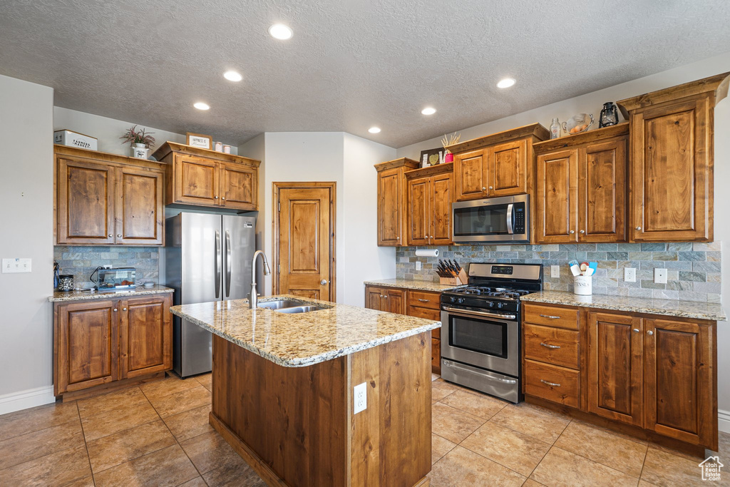 Kitchen with stainless steel appliances, brown cabinets, a sink, backsplash, and an island with sink