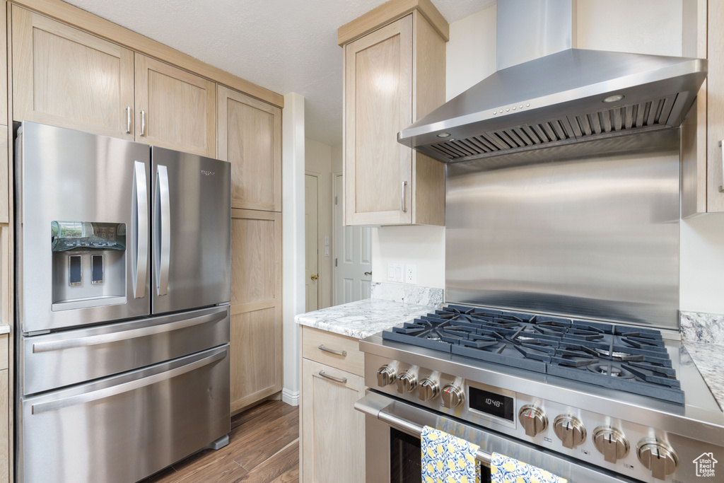 Kitchen featuring stainless steel appliances, wall chimney range hood, light brown cabinetry, wood finished floors, and light stone countertops