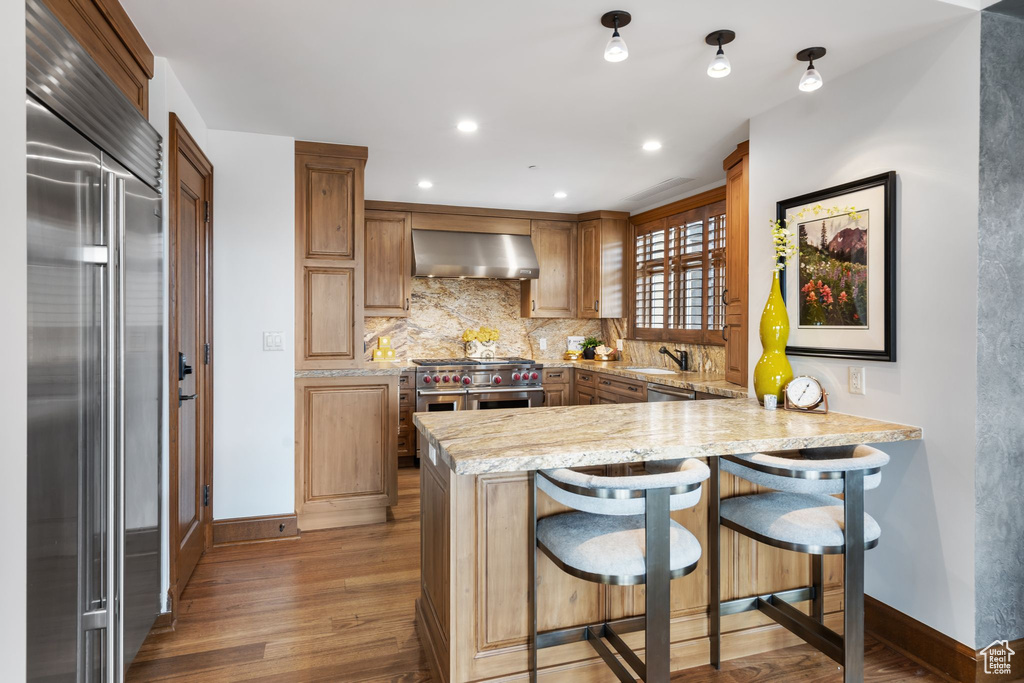 Kitchen with high quality appliances, wall chimney range hood, light wood finished floors, a peninsula, and recessed lighting