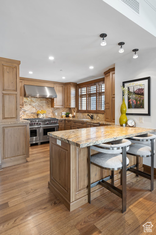 Kitchen with wall chimney range hood, appliances with stainless steel finishes, decorative backsplash, a kitchen bar, and light wood-style floors