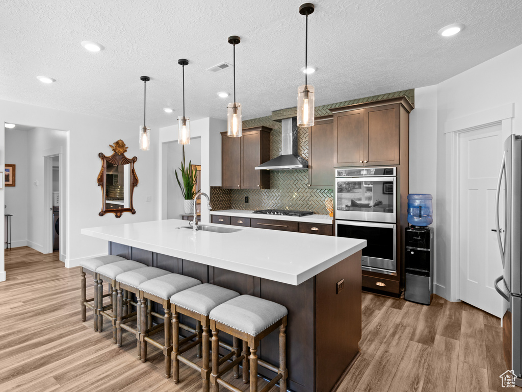 Kitchen featuring a sink, stainless steel appliances, wall chimney range hood, light wood-style floors, and a textured ceiling