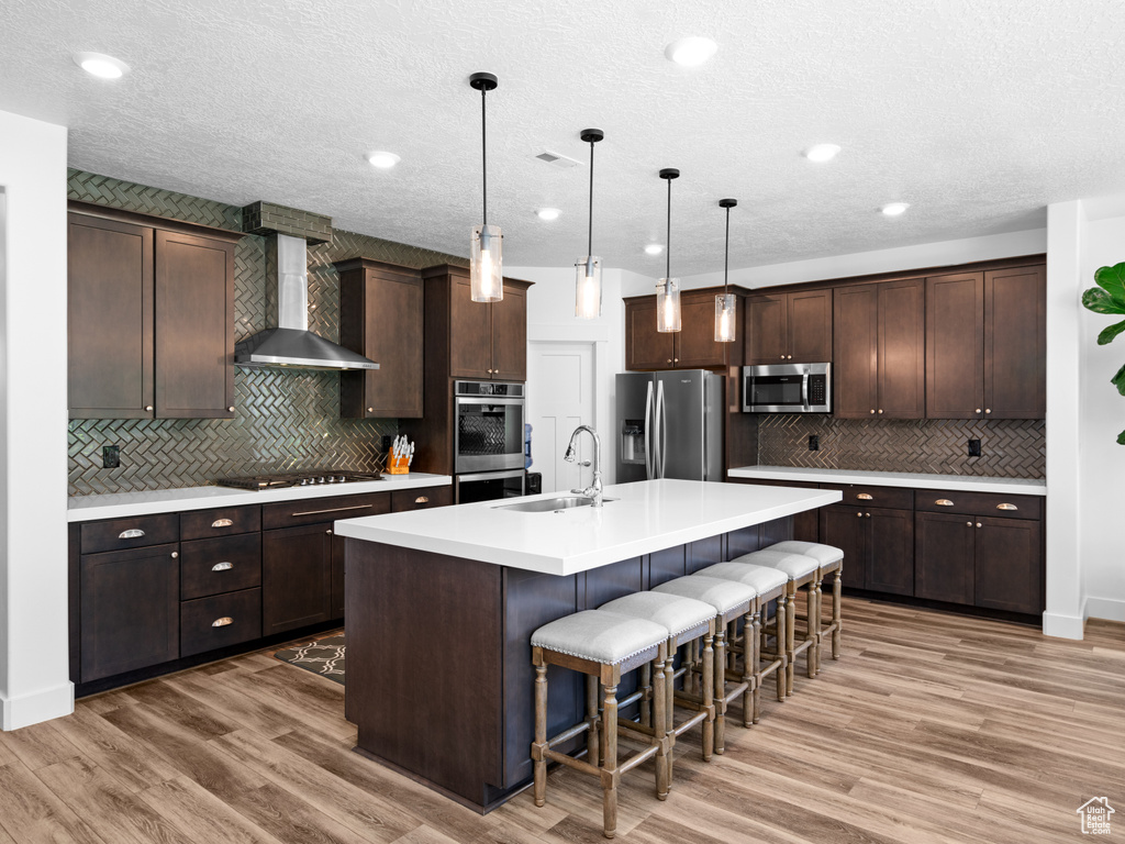 Kitchen featuring appliances with stainless steel finishes, a sink, wall chimney range hood, decorative backsplash, and dark brown cabinetry