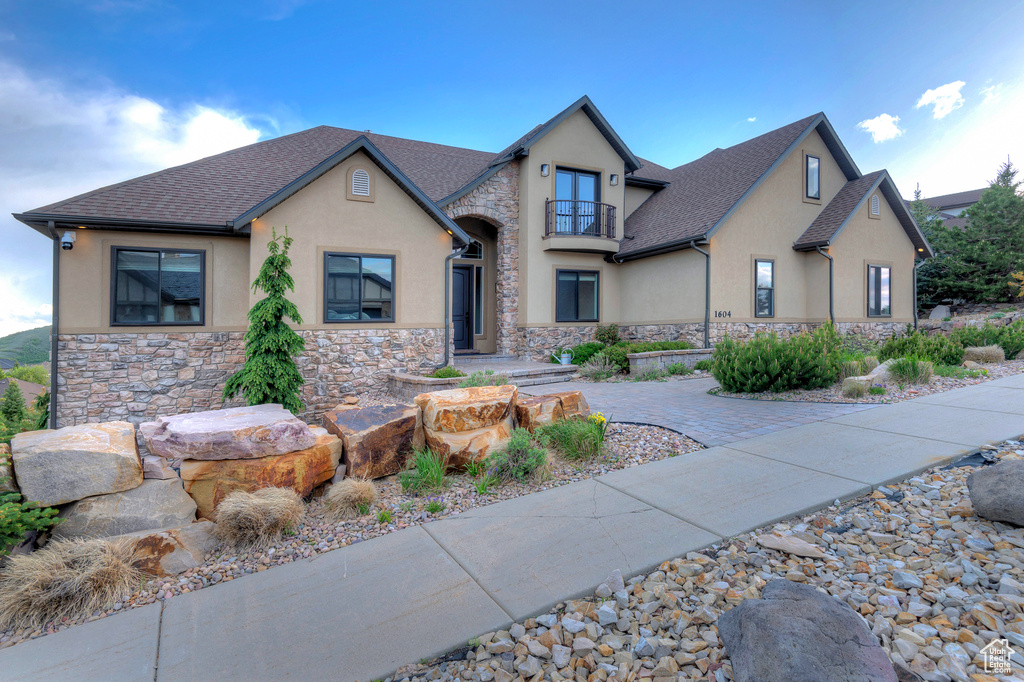French country home with a balcony, stucco siding, stone siding, and roof with shingles