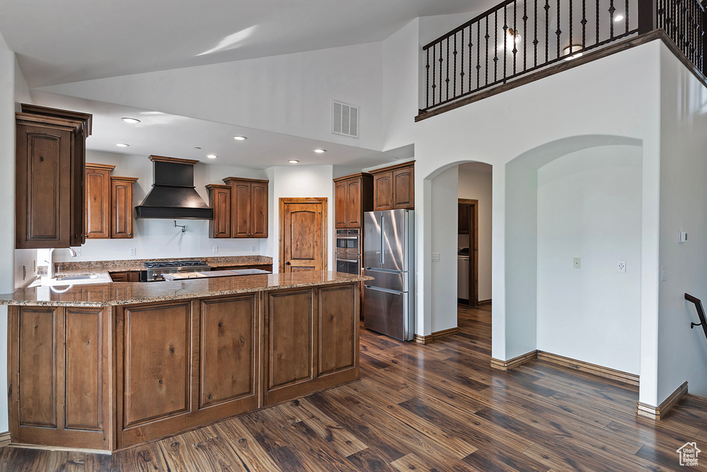 Kitchen with custom exhaust hood, stainless steel appliances, dark wood-style floors, arched walkways, and a peninsula
