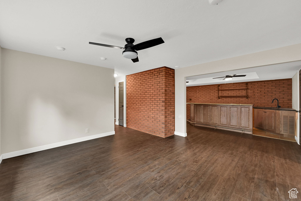 Unfurnished living room featuring ceiling fan, brick wall, dark wood finished floors, a sink, and baseboards