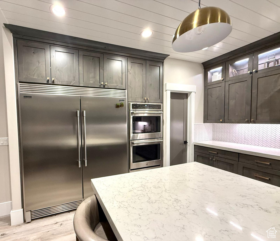 Kitchen with stainless steel appliances, light wood-type flooring, tasteful backsplash, light stone countertops, and decorative light fixtures