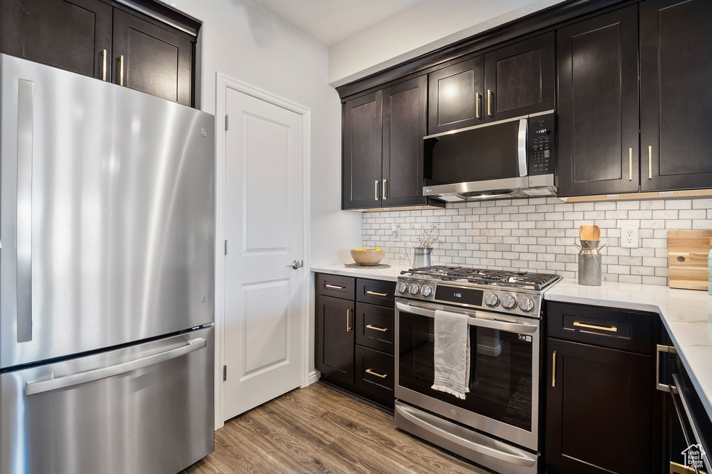 Kitchen with stainless steel appliances, decorative backsplash, wood finished floors, dark brown cabinetry, and light stone countertops
