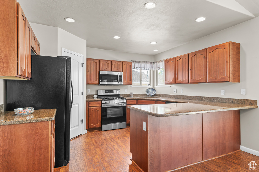 Kitchen featuring stainless steel appliances, a peninsula, wood finished floors, brown cabinetry, and recessed lighting