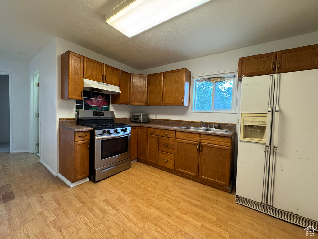 Kitchen with white refrigerator with ice dispenser, stainless steel gas range, under cabinet range hood, and brown cabinetry