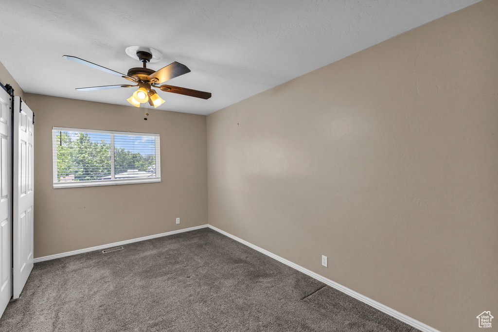 Empty room with ceiling fan, a barn door, baseboards, and dark carpet