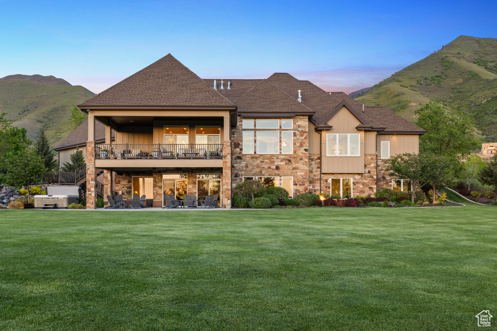 Back of house at dusk featuring a lawn, stone siding, a shingled roof, and a mountain view