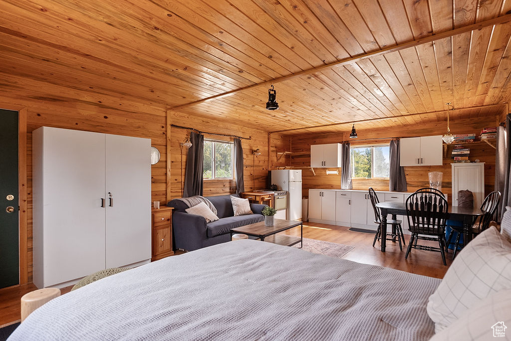 Bedroom with multiple windows, wooden ceiling, light wood-style flooring, freestanding refrigerator, and rustic walls
