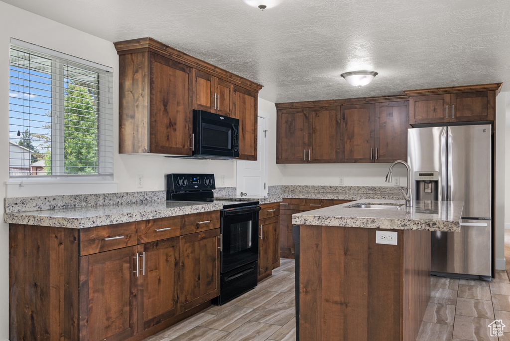 Kitchen featuring black appliances, a sink, a kitchen island with sink, light stone counters, and a textured ceiling