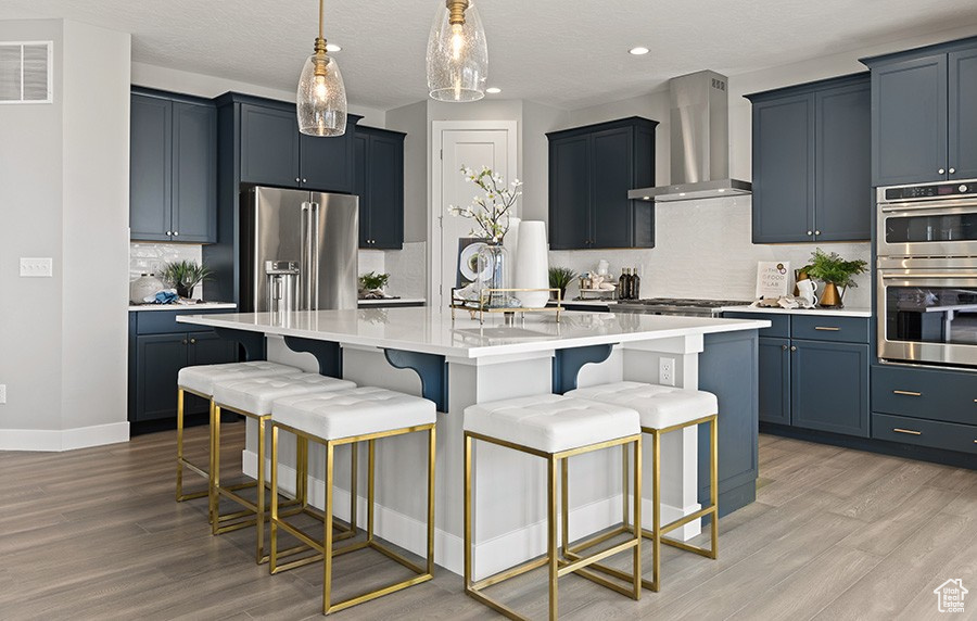 Kitchen with wall chimney range hood, stainless steel appliances, light wood finished floors, tasteful backsplash, and a center island with sink