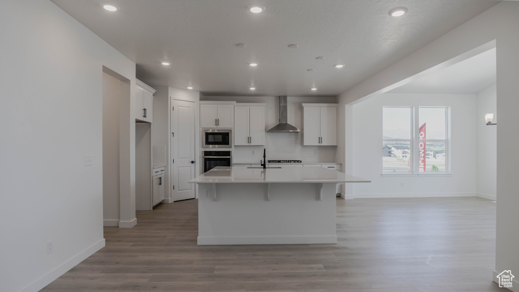 Kitchen featuring wall chimney range hood, black microwave, stainless steel oven, a kitchen island with sink, and white cabinetry