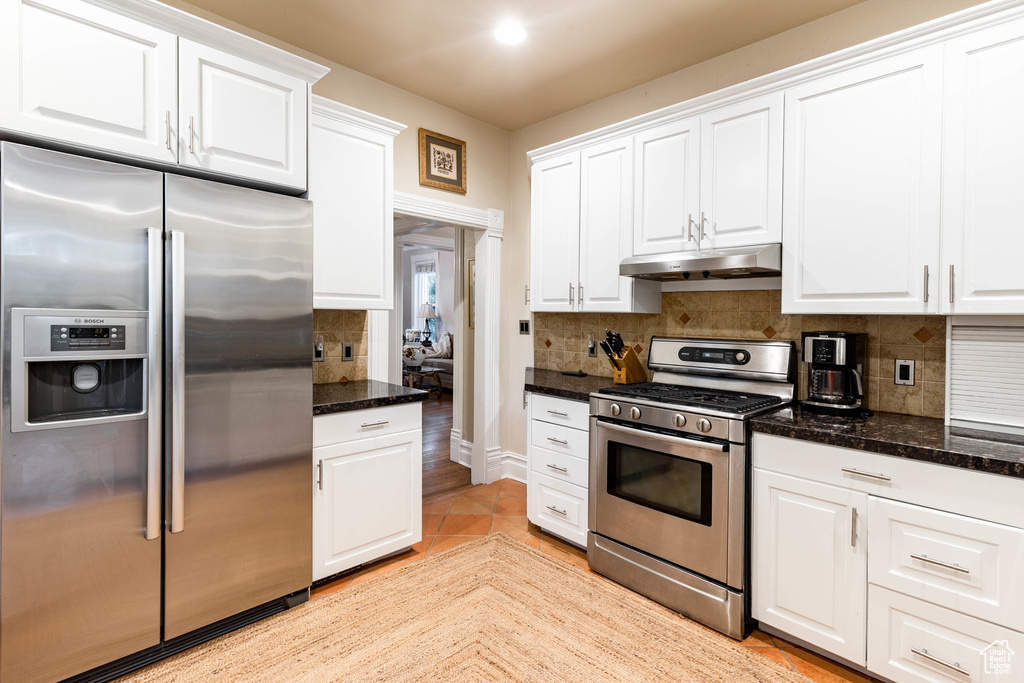 Kitchen with appliances with stainless steel finishes, under cabinet range hood, white cabinets, tasteful backsplash, and dark stone counters