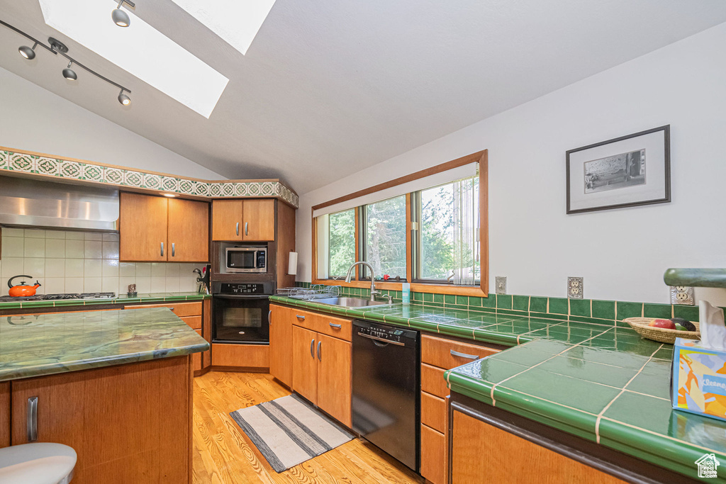 Kitchen featuring black appliances, a sink, a skylight, wall chimney range hood, and vaulted ceiling