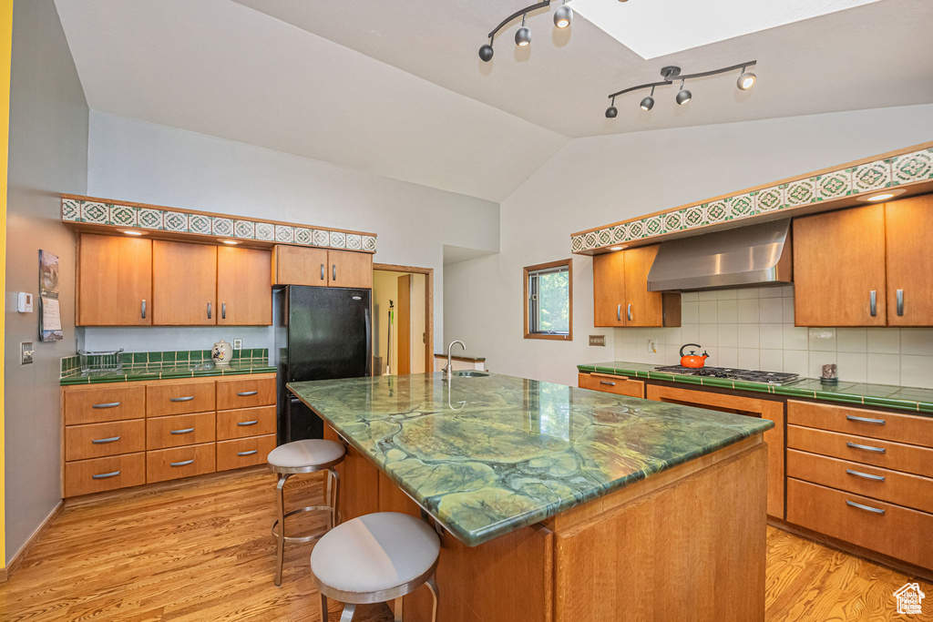 Kitchen featuring wall chimney range hood, stainless steel gas stovetop, light wood finished floors, vaulted ceiling, and a skylight