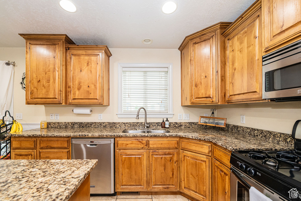 Kitchen with stainless steel appliances, a sink, dark stone counters, brown cabinetry, and recessed lighting
