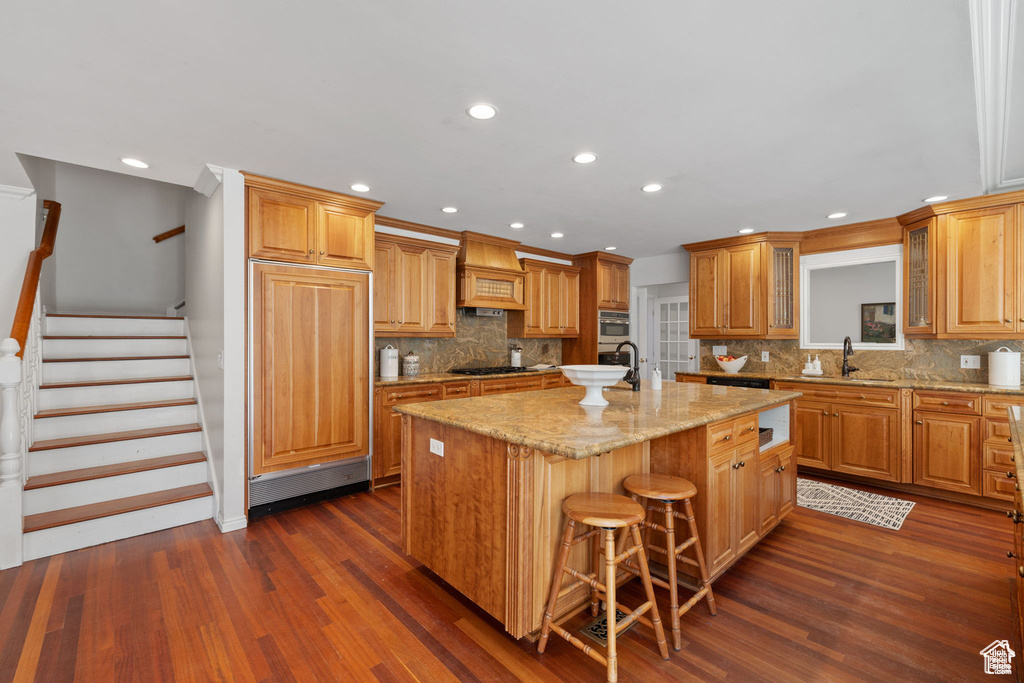 Kitchen featuring premium range hood, a center island with sink, black gas cooktop, dark wood-style floors, and tasteful backsplash