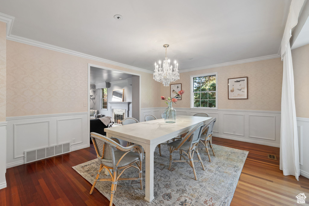 Dining space featuring wallpapered walls, crown molding, a warm lit fireplace, a chandelier, and wood finished floors