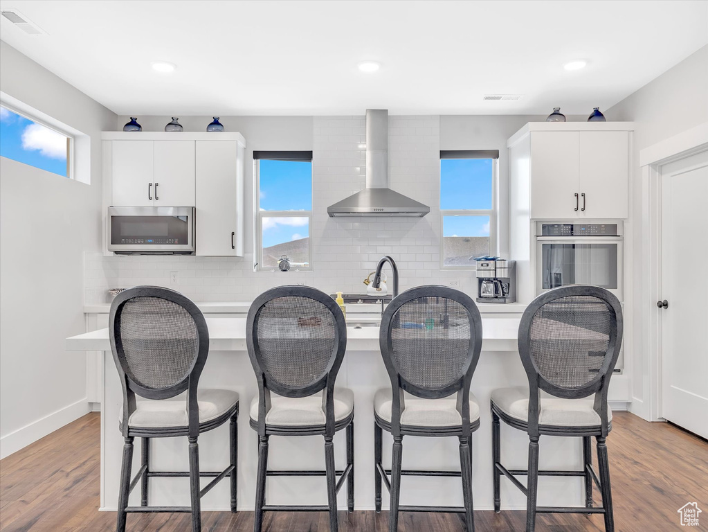 Kitchen with appliances with stainless steel finishes, wall chimney range hood, a kitchen island with sink, backsplash, and a kitchen breakfast bar
