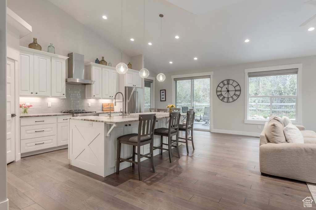 Kitchen featuring wall chimney range hood, an island with sink, tasteful backsplash, white cabinets, and recessed lighting