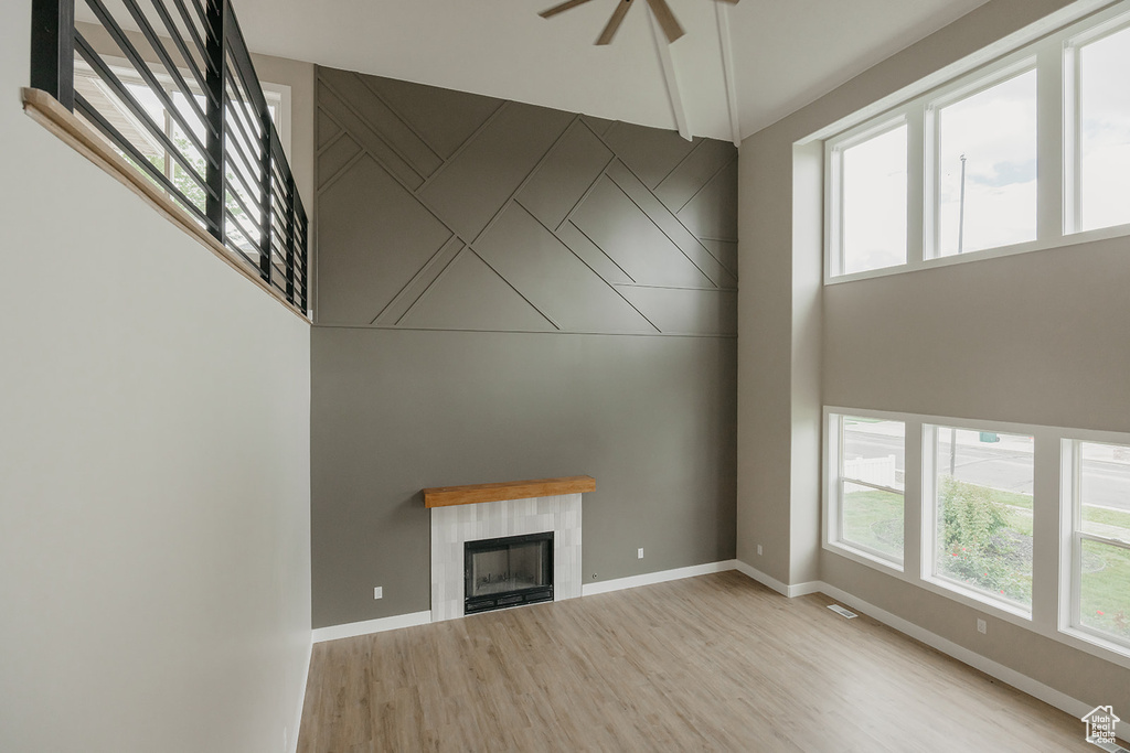 Unfurnished living room featuring light wood-type flooring, plenty of natural light, ceiling fan, a tiled fireplace, and a high ceiling