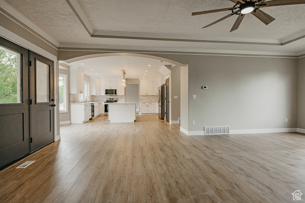 Unfurnished living room featuring a tray ceiling, a textured ceiling, arched walkways, a ceiling fan, and light wood-style flooring
