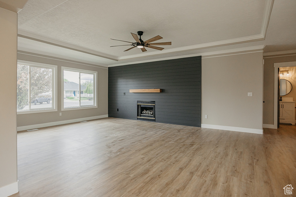 Unfurnished living room featuring a tray ceiling, a ceiling fan, light wood-style floors, crown molding, and a large fireplace