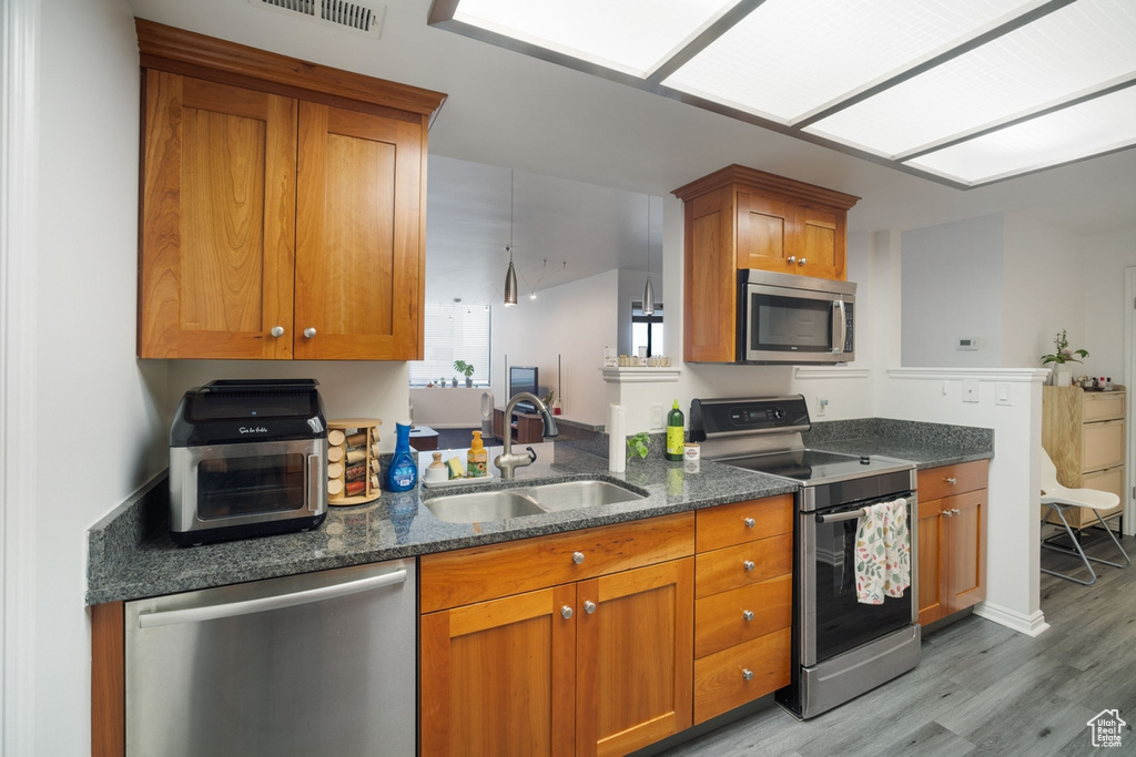 Kitchen featuring appliances with stainless steel finishes, a sink, brown cabinetry, light wood-style flooring, and dark stone counters