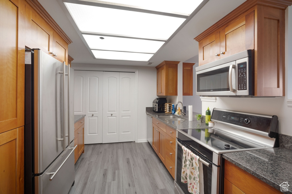 Kitchen with stainless steel appliances, a sink, light wood-type flooring, brown cabinets, and dark stone counters