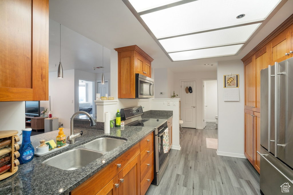 Kitchen with appliances with stainless steel finishes, a sink, brown cabinetry, light wood-type flooring, and dark stone counters