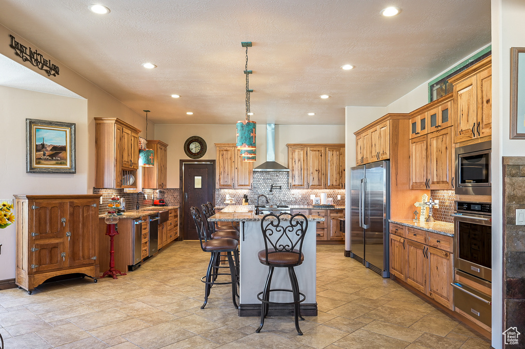 Kitchen with stainless steel appliances, wall chimney exhaust hood, an island with sink, a warming drawer, and light stone countertops