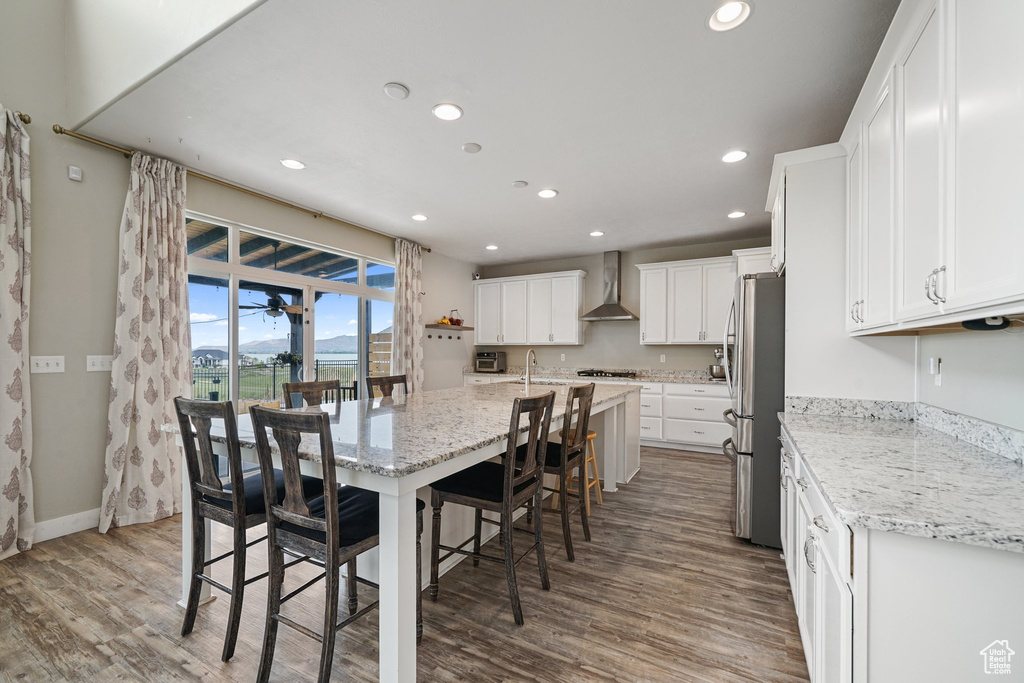 Kitchen featuring wall chimney exhaust hood, stainless steel appliances, recessed lighting, wood finished floors, and a kitchen bar