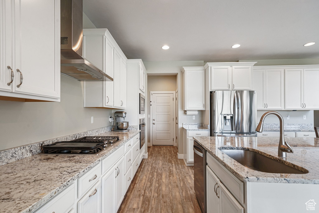 Kitchen with stainless steel appliances, a sink, wall chimney range hood, wood finished floors, and white cabinets