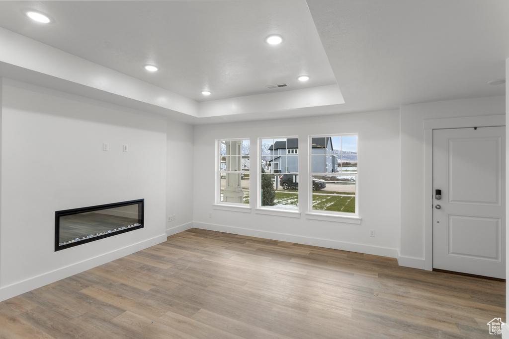 Unfurnished living room featuring a raised ceiling, baseboards, a glass covered fireplace, light wood-style flooring, and recessed lighting
