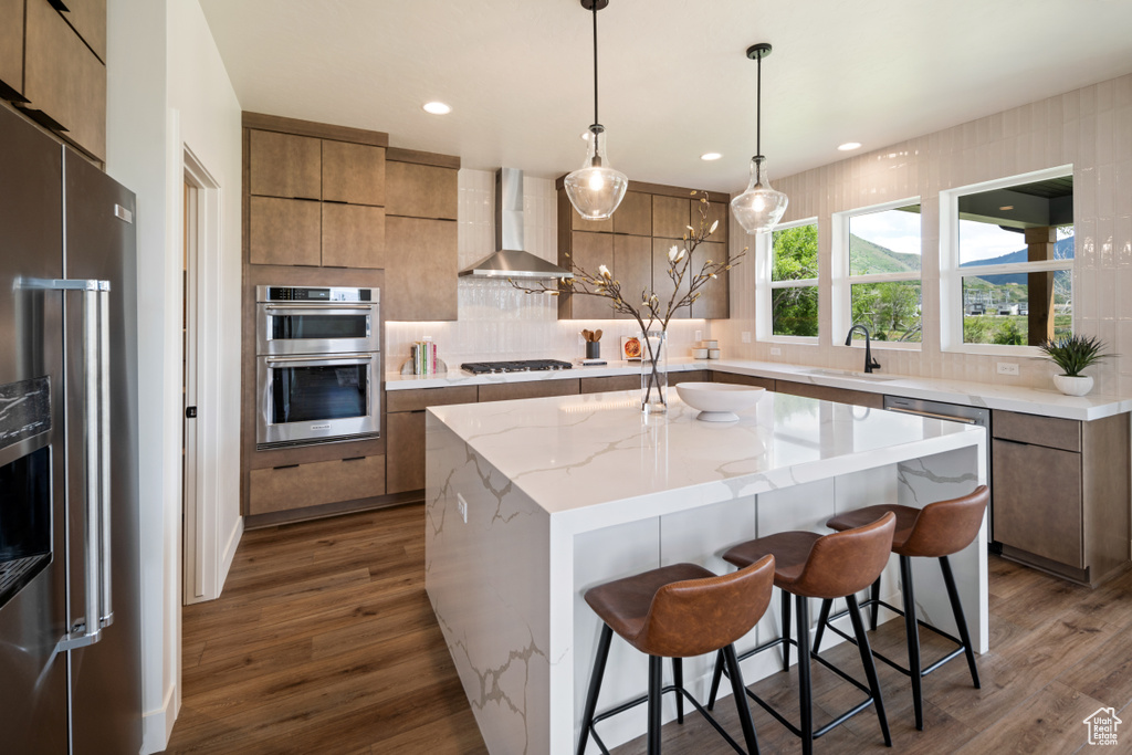 Kitchen with stainless steel appliances, wall chimney range hood, a sink, dark wood finished floors, and a kitchen island