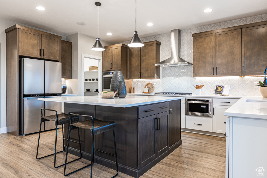 Kitchen with appliances with stainless steel finishes, wall chimney range hood, light wood-type flooring, a center island, and recessed lighting