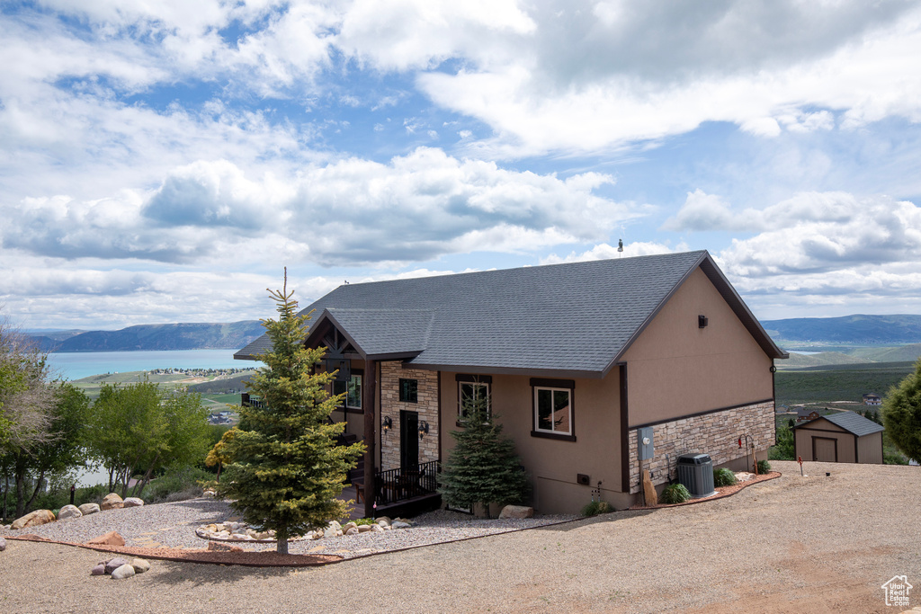 View of front of house with stone siding, central air condition unit, a water and mountain view, an outbuilding, and a shingled roof