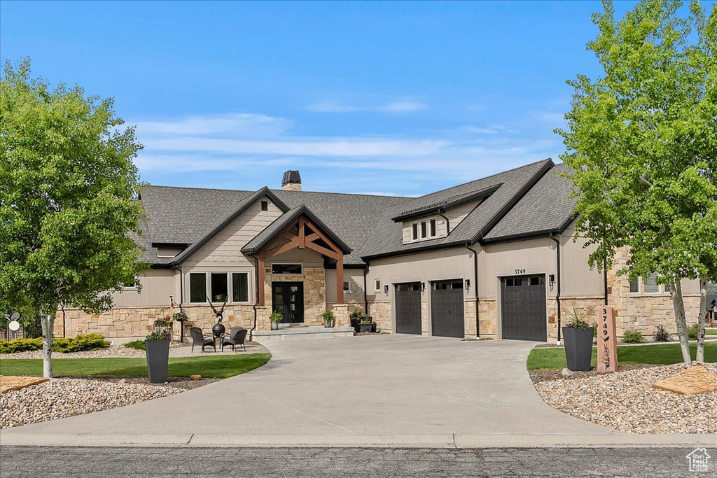 View of front of home with driveway, stone siding, a shingled roof, a chimney, and a garage