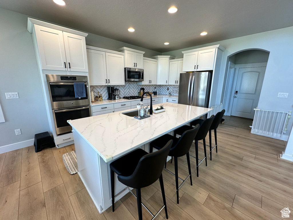 Kitchen featuring stainless steel appliances, a sink, a kitchen island with sink, white cabinetry, and light stone counters