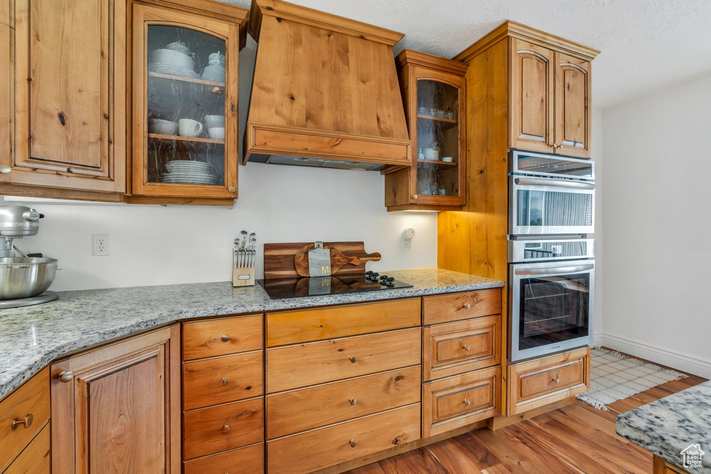 Kitchen with custom range hood, light stone counters, black electric stovetop, glass insert cabinets, and light wood-style floors
